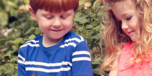 Two children, a boy with red hair and a girl with curly hair, are gently holding butterflies on their hands while smiling. They are surrounded by lush greenery and colorful flowers, enjoying a moment in nature.