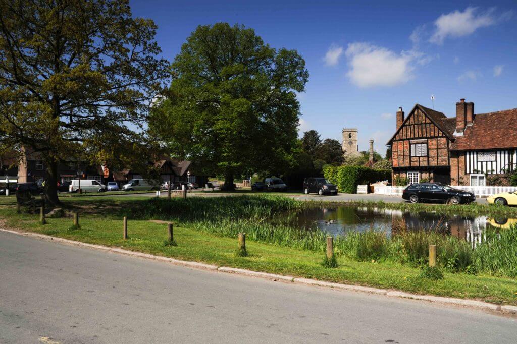 A picturesque scene of a village with a pond surrounded by greenery. Traditional-style houses and a church tower are visible in the background, under a clear blue sky. Cars are parked along the road, enhancing the quaint charm of the setting.