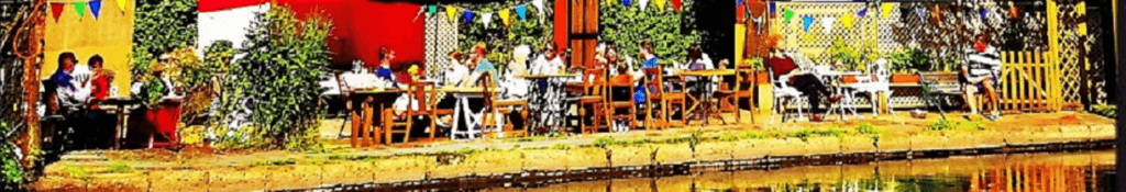 A vibrant outdoor café scene with several people sitting at tables along a waterway, surrounded by greenery and colorful decorations, creating a lively and inviting atmosphere.