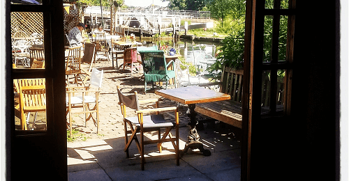 A view from an open doorway overlooking a quaint outdoor café area with tables and chairs beside a body of water, surrounded by greenery and umbrellas.