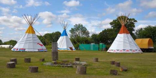 Three colorful tipis sit in a grassy field under a partly cloudy sky, surrounded by a circular arrangement of stones. In the background, additional structures are visible, creating a peaceful outdoor setting.