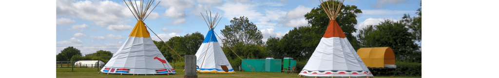A series of traditional tepees are displayed in a grassy area, with trees and a blue sky in the background. Each tepee features distinct colors on top, including orange, blue, and red, indicating a vibrant, cultural setting.