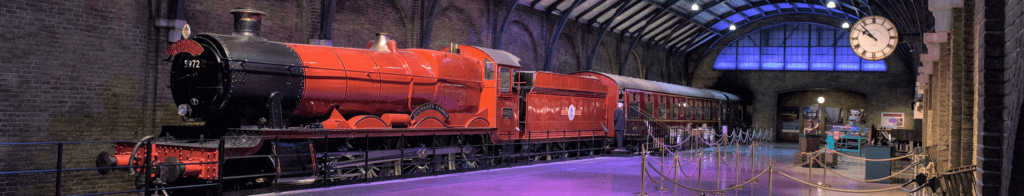 A vibrant red steam locomotive is displayed in a dimly lit train station, featuring historic architecture. The train is positioned on tracks, with a clock visible on the wall, emphasizing the nostalgic atmosphere of the scene.