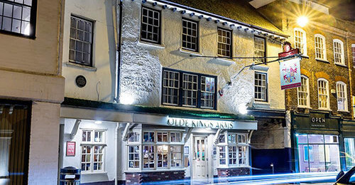 A historic building at night, featuring a pub with a sign and warm lighting. The structure has multiple windows and a decorative exterior, surrounded by other shops. Subtle light trails hint at passing traffic.