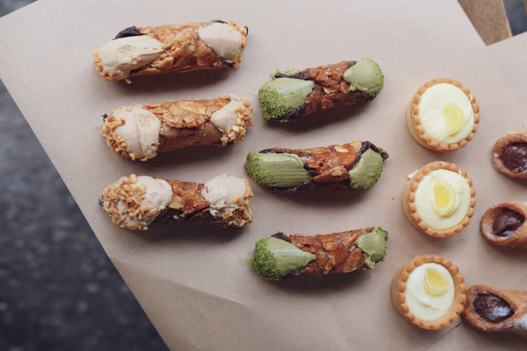 An assortment of desserts including three types of cannoli filled with cream, some coated with nuts or pistachio, and mini custard tarts arranged on a sheet of parchment paper.