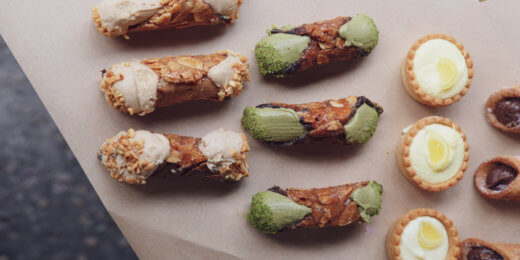 An assortment of desserts including three types of cannoli filled with cream, some coated with nuts or pistachio, and mini custard tarts arranged on a sheet of parchment paper.