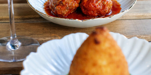 Two dishes on white plates: one features meatballs in a rich tomato sauce, and the other presents a crispy, conical fritter on a bed of green sauce, accompanied by a glass of white wine.