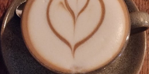 A latte in a green cup, featuring a heart-shaped foam art design on top, sits on a textured wooden surface.