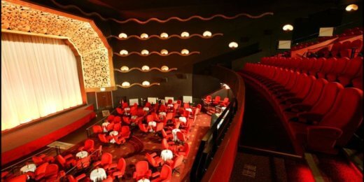 An elegant theater interior featuring plush red seating arranged around tables with white tablecloths, illuminated by soft lighting. A large stage with a decorative backdrop is in the front, creating a sophisticated atmosphere suitable for performances or events.
