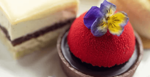 An assortment of elegant desserts, including a vibrant red dome-shaped cake topped with a purple flower, alongside two squares of layered cake with cream and chocolate filling, displayed on a white plate.