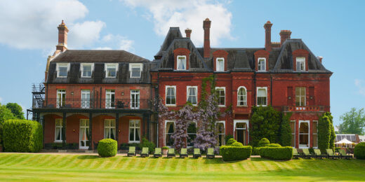 A grand red-brick mansion with intricate architectural details, surrounded by neatly trimmed green lawns and topiary. The sky is clear, enhancing the elegance of the estate.