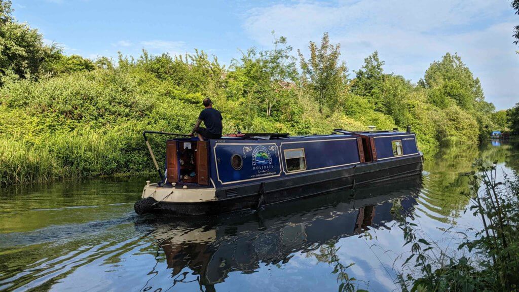 A narrowboat glides smoothly along a tranquil waterway, surrounded by lush greenery. A person stands at the helm, navigating the boat under a clear blue sky.