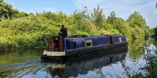 A narrowboat glides smoothly along a tranquil waterway, surrounded by lush greenery. A person stands at the helm, navigating the boat under a clear blue sky.