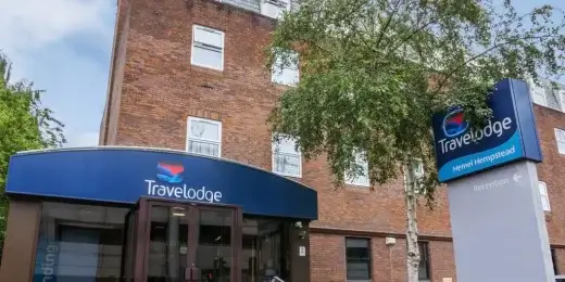 A Travelodge hotel building with a blue roof and signage. The entrance features steps leading up to glass doors, surrounded by greenery. The façade of the building is brick, and a large sign displays the Travelodge logo.