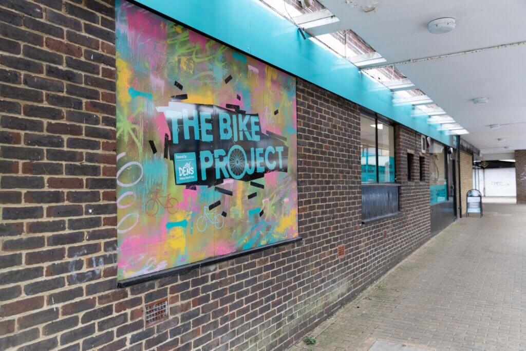 A mural featuring colorful graffiti artwork on a brick wall advertises "The Bike Project." The vibrant design includes bold text and abstract patterns, set against a backdrop of a covered walkway with overhead lighting.