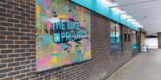 A mural featuring colorful graffiti artwork on a brick wall advertises "The Bike Project." The vibrant design includes bold text and abstract patterns, set against a backdrop of a covered walkway with overhead lighting.