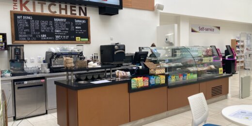 A café counter named "The Kitchen" featuring a menu board, coffee machines, and a display case filled with snacks and beverages. Brightly lit with modern decor, the setting encourages casual dining.