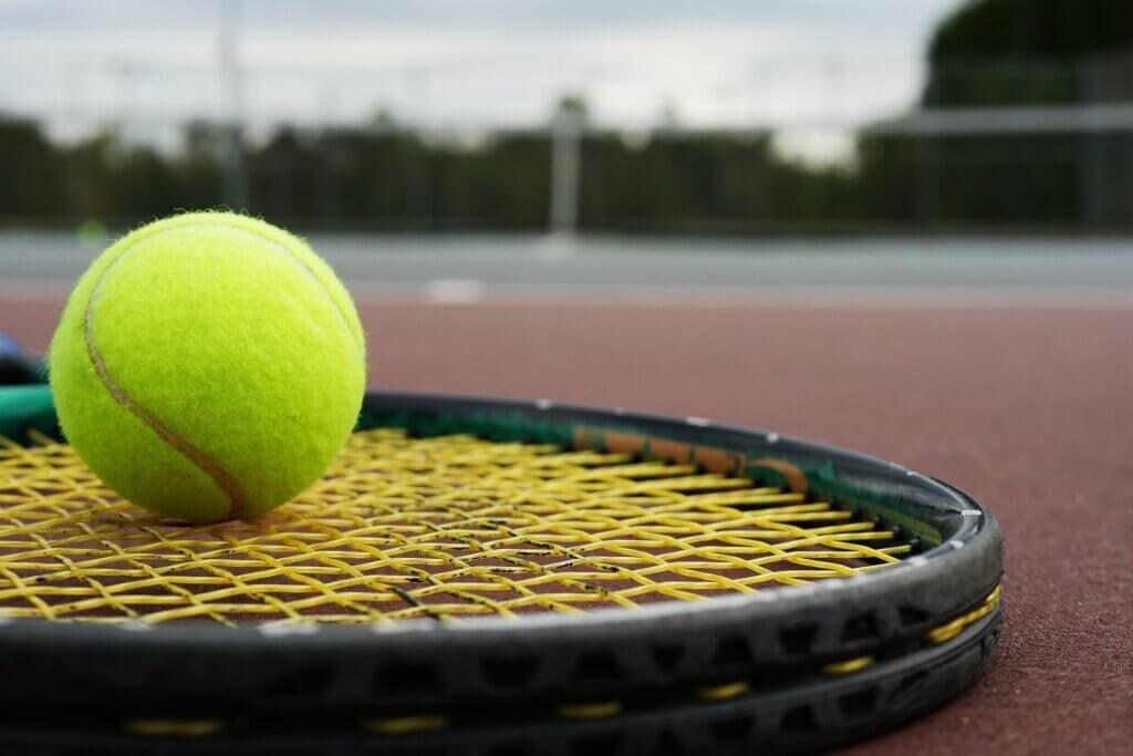 A close-up view of a bright yellow tennis ball resting on a black and yellow tennis racket, with a blurred tennis court background.