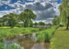 A serene riverside scene featuring a gently flowing stream bordered by lush greenery and trees. Dramatic clouds fill the sky, creating a picturesque landscape perfect for nature lovers.