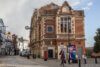 A charming brick building with decorative features stands at the corner of a street. A woman walks by, and a colorful owl sculpture is visible nearby. The scene captures a lively urban atmosphere under a partly cloudy sky.