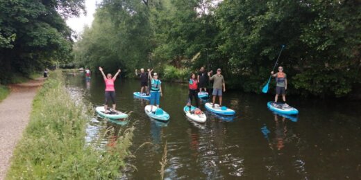 A group of people paddleboarding on a calm river surrounded by lush greenery. Some participants are standing and cheering while others are paddling on blue boards. A path runs alongside the water, enhancing the outdoor activity atmosphere.