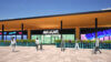 An outdoor view of a modern building featuring the sign "Mulligans." Pedestrians walk along a paved area in front, with colorful planters lining the entrance. The sky is clear and sunny, enhancing the vibrant atmosphere of the location.