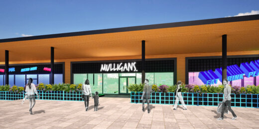 An outdoor view of a modern building featuring the sign "Mulligans." Pedestrians walk along a paved area in front, with colorful planters lining the entrance. The sky is clear and sunny, enhancing the vibrant atmosphere of the location.