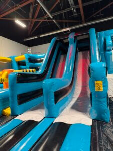 Large indoor inflatable slide with bright blue side rails, leading down to a padded base. Overhead lights and exposed ceiling beams are visible in the background, suggesting an indoor play area.