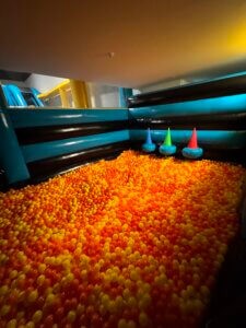 A large indoor ball pit filled with orange and yellow balls, surrounded by inflatable black and teal barriers. Three colorful cone-shaped objects are placed against the back wall.