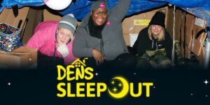 Three smiling people in warm clothing inside a cardboard shelter, participating in the DENS sleepout event.